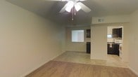 This single-story space features an open-concept layout that connects the living area to the kitchen, creating a seamless flow throughout the unit. The living room is finished with wall-to-wall carpeting in a neutral tone, contributing to a warm and inviting atmosphere. A ceiling fan with an integrated light fixture is centrally mounted, providing both air circulation and ambient lighting. The walls are painted in a soft, neutral hue, complementing the understated aesthetic of the space. A window positioned toward the rear of the living area allows natural light to filter in, brightening the interior. The kitchen is partially visible from the living room and features dark cabinetry, offering a subtle contrast against the lighter surroundings. A tile or hard-surface floor transitions into the kitchen zone, distinguishing the two areas. A HVAC vent is visible in the ceiling, indicating a central air system throughout. The overall design is clean and functional, with straightforward finishes suited for comfortable everyday living.