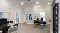 This single-story professional office space is thoughtfully designed to convey both sophistication and functionality. The main office area features notably high ceilings accented with classic crown molding, lending the space an open and refined character. Neutral grey and beige tones throughout the walls and carpeted flooring create a calm, professional ambiance.

The primary workspace is anchored by a sleek glass-top desk accompanied by modern chrome-framed chairs, offering a contemporary contrast to the traditionally styled architectural details. Framed credentials and artwork are displayed on the walls, reinforcing a professional and credentialed image. An arched window allows natural light to filter into the space, brightening the interior while maintaining privacy.

Multiple doorways lead to adjoining rooms, with one featuring a glass-paneled door that hints at additional private office or meeting space beyond. Recessed lighting throughout provides consistent, well-distributed illumination. The overall design balances classic architectural elements with modern furnishings, resulting in a polished environment well-suited for professional consultations and client interactions.