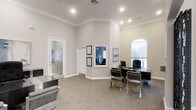 This single-story professional office space is thoughtfully designed to convey both sophistication and functionality. The main office area features notably high ceilings accented with classic crown molding, lending the space an open and refined character. Neutral grey and beige tones throughout the walls and carpeted flooring create a calm, professional ambiance.

The primary workspace is anchored by a sleek glass-top desk accompanied by modern chrome-framed chairs, offering a contemporary contrast to the traditionally styled architectural details. Framed credentials and artwork are displayed on the walls, reinforcing a professional and credentialed image. An arched window allows natural light to filter into the space, brightening the interior while maintaining privacy.

Multiple doorways lead to adjoining rooms, with one featuring a glass-paneled door that hints at additional private office or meeting space beyond. Recessed lighting throughout provides consistent, well-distributed illumination. The overall design balances classic architectural elements with modern furnishings, resulting in a polished environment well-suited for professional consultations and client interactions.