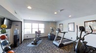 This fitness room offers a practical and well-organized space designed to support cardiovascular exercise and general wellness. The room features a patterned carpet flooring that adds visual texture while providing a comfortable surface underfoot. Recessed lighting in the textured ceiling ensures the space is evenly and adequately lit throughout. A window fitted with horizontal blinds along one wall allows natural light to enter, contributing to an open and welcoming atmosphere. The cardio equipment on offer includes a treadmill, an elliptical machine, and a recumbent stationary bike, each positioned with ample spacing to allow for safe and comfortable use. Rubber exercise mats beneath the equipment help protect the flooring and reduce noise during workouts. Informational charts and notices are mounted on the walls, providing users with reference materials during their sessions. The neutral white walls keep the environment feeling clean and focused. Overall, the space is thoughtfully arranged to maximize functionality within its footprint, making it a convenient and accessible fitness option.