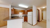 This single-floor residence offers a well-organized and functional layout designed to maximize everyday comfort. The kitchen serves as a central highlight, featuring warm-toned wood cabinetry that spans both upper and lower sections, providing generous storage throughout. A peninsula counter extends into the space, offering additional prep area and a natural division between the kitchen and adjoining areas. White appliances, including a full-size refrigerator and electric range with an overhead ventilation hood, are neatly integrated into the layout. Bright overhead flush lighting illuminates the workspace effectively, while a pendant light adds a subtle decorative touch near the peninsula. Light-toned flooring anchors the kitchen, contributing to a clean and open feel. A hallway visible from the kitchen suggests a straightforward and accessible floor plan, connecting the main living areas with ease. The overall ambiance is practical and unpretentious, with neutral tones and classic finishes creating a welcoming, move-in-ready environment suited for comfortable daily living.