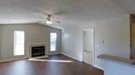 This single-story space presents a well-designed living area that serves as the central gathering point of the home. The room features vaulted ceilings that enhance the sense of openness, complemented by a ceiling fan with integrated lighting for both comfort and ambiance. Dark hardwood flooring runs throughout the main living space, providing a warm contrast against the neutral gray walls and white trim.

A tiled fireplace anchors one wall of the living area, adding a focal point and a cozy element to the room. Natural light filters in through a window positioned near the fireplace, brightening the space during daylight hours. A ceiling vent indicates a central HVAC system for year-round climate control.

An open doorway leads to an adjacent room with carpeted flooring, suggesting a bedroom or secondary living space. A partial view of what appears to be a countertop along the right edge of the image hints at a nearby kitchen or bar area. The textured ceiling and clean, minimal design give the space a straightforward and functional character, suitable for a variety of living arrangements.