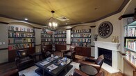 This refined library lounge offers a warm, scholarly atmosphere defined by its thoughtful architectural details and carefully curated furnishings. Built-in bookshelves line nearly every wall, housed within arched alcoves framed by decorative columns and dark wood trim, and stocked with an extensive collection of books spanning multiple genres. A classic white-surround fireplace serves as a focal point on one wall, flanked by additional shelving and accented by a large vintage-style clock, adding to the room's distinguished character. Overhead, a striking orb-style chandelier with exposed Edison-style bulbs casts warm, ambient light across the space, complemented by recessed ceiling lighting. The ceiling features a rich golden-amber tone that enhances the cozy, intimate feel of the room. Seating consists of multiple deep brown leather armchairs arranged around low glass-topped coffee tables displaying an assortment of magazines and reading materials, inviting guests to settle in comfortably. The flooring is rich hardwood, partially covered by a neutral area rug that anchors the central seating arrangement. French doors provide access to an adjacent space and allow natural light to filter in through slatted blinds. The overall ambiance is elegant yet approachable, making this library a welcoming retreat for quiet reading, conversation, or leisure.