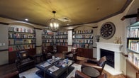 This refined library lounge offers a warm, scholarly atmosphere defined by its thoughtful architectural details and carefully curated furnishings. Built-in bookshelves line nearly every wall, housed within arched alcoves framed by decorative columns and dark wood trim, and stocked with an extensive collection of books spanning multiple genres. A classic white-surround fireplace serves as a focal point on one wall, flanked by additional shelving and accented by a large vintage-style clock, adding to the room's distinguished character. Overhead, a striking orb-style chandelier with exposed Edison-style bulbs casts warm, ambient light across the space, complemented by recessed ceiling lighting. The ceiling features a rich golden-amber tone that enhances the cozy, intimate feel of the room. Seating consists of multiple deep brown leather armchairs arranged around low glass-topped coffee tables displaying an assortment of magazines and reading materials, inviting guests to settle in comfortably. The flooring is rich hardwood, partially covered by a neutral area rug that anchors the central seating arrangement. French doors provide access to an adjacent space and allow natural light to filter in through slatted blinds. The overall ambiance is elegant yet approachable, making this library a welcoming retreat for quiet reading, conversation, or leisure.