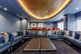 This sophisticated single-floor lounge and media space showcases a striking interior design centered around a bold circular cove ceiling with warm ambient uplighting, set against deep teal and slate-blue walls that create an immersive, upscale atmosphere throughout.

The main lounge area is anchored by a generously sized curved sectional sofa in dark leather and fabric, accented with decorative pillows in earth tones and geometric patterns. Large leather ottomans provide additional flexible seating and surface space, while patterned carpet in muted tones grounds the room with texture and depth.

At one end of the space, a projection screen is mounted on the wall, oriented toward the seating arrangement, making this area well-suited for private screenings or presentations. Recessed ceiling lighting complements the cove's ambient glow, ensuring even illumination throughout.

The adjacent lounge and bar area features a sleek built-in wet bar with dark wood cabinetry and a countertop sink, flanked by framed artwork on the walls. Bold red-orange bar stools surround high-top tables positioned near large windows, which frame elevated city views and allow natural light to filter through dramatic floor-length drapes. The combination of curated furnishings, architectural lighting, and city vistas makes this a highly functional and visually compelling entertainment venue.