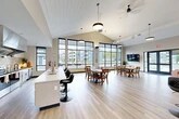 Located at 22 Loso Dr in Cicero, NY, this single-floor community clubhouse offers a generous, open-concept layout designed for gathering and entertaining. The space is anchored by soaring vaulted ceilings with a shiplap-style finish and floor-to-ceiling grid-pane windows that flood the interior with natural light and frame sweeping views of the surrounding outdoor patio and greenery.

The main area is thoughtfully organized into distinct functional zones. A casual lounge features a plush tufted sofa and leather accent chairs arranged around a flat-screen TV, opening directly to the outdoor patio through large glass doors. High-top bistro tables with upholstered bar-height chairs provide additional seating for socializing. A traditional dining area accommodates multiple tables with wooden chairs, ideal for group meals or events.

The full-service kitchen is outfitted with stainless steel appliances, including a full-size refrigerator, range with a commercial-grade hood, dishwasher, and a spacious white quartz island with bar seating. Neutral tile backsplash and white cabinetry complete the clean, modern aesthetic. Light wood-look flooring runs throughout the entire space, tying the zones together seamlessly. Pendant lighting and ceiling fans contribute to the bright, welcoming ambiance of this well-appointed communal facility.