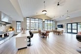 Located at 22 Loso Dr in Cicero, NY, this single-floor community clubhouse offers a generous, open-concept layout designed for gathering and entertaining. The space is anchored by soaring vaulted ceilings with a shiplap-style finish and floor-to-ceiling grid-pane windows that flood the interior with natural light and frame sweeping views of the surrounding outdoor patio and greenery.

The main area is thoughtfully organized into distinct functional zones. A casual lounge features a plush tufted sofa and leather accent chairs arranged around a flat-screen TV, opening directly to the outdoor patio through large glass doors. High-top bistro tables with upholstered bar-height chairs provide additional seating for socializing. A traditional dining area accommodates multiple tables with wooden chairs, ideal for group meals or events.

The full-service kitchen is outfitted with stainless steel appliances, including a full-size refrigerator, range with a commercial-grade hood, dishwasher, and a spacious white quartz island with bar seating. Neutral tile backsplash and white cabinetry complete the clean, modern aesthetic. Light wood-look flooring runs throughout the entire space, tying the zones together seamlessly. Pendant lighting and ceiling fans contribute to the bright, welcoming ambiance of this well-appointed communal facility.