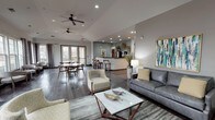 This single-floor space is designed as a welcoming communal gathering area, combining multiple functional zones within an open, flowing layout. The interior is anchored by a dramatic floor-to-ceiling stacked stone fireplace, which serves as a prominent architectural focal point. Vaulted ceilings with recessed lighting and ceiling fans add to the sense of openness, while dark hardwood flooring runs throughout, providing a warm and cohesive foundation.

The lounge area features a gray leather sofa, upholstered accent chairs, and a marble-topped coffee table arranged on a patterned area rug, complemented by colorful abstract artwork on the walls. A wall-mounted television sits above a wooden console near the fireplace. Large French doors and floor-to-ceiling windows line the perimeter, flooding the space with natural light and offering views of the surrounding exterior.

A dedicated dining area includes a round marble-top table with mid-century modern wooden chairs, positioned near sliding glass doors. The kitchen features stainless steel appliances—including a French door refrigerator and gas range—dark cabinetry, black countertops, and a bar-height counter with seating. Decorative woven wall accents and greenery complete the polished, contemporary aesthetic throughout.