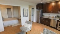 This single-floor residence offers an open, functional layout that connects the main living area directly to the kitchen, creating a cohesive and easy-to-navigate space. Light hardwood-style flooring runs throughout, lending warmth and visual continuity across the unit.

The kitchen is outfitted with rich dark-brown cabinetry, light stone-toned countertops, and stainless steel appliances, including a full-sized refrigerator. Track lighting overhead ensures the kitchen remains well-lit and practical for everyday use. A dedicated in-unit laundry closet is conveniently positioned nearby, housing a stacked washer and dryer behind louvered doors.

The living area provides enough room for comfortable seating arrangements, as seen with the patterned accent chairs and matching ottoman. Wall art adds a decorative focal point to the otherwise clean, neutral wall space. A separate bedroom is accessible through a doorway off the main living area, offering a degree of privacy. The bedroom appears carpeted, providing a softer feel distinct from the rest of the unit.

Overall, the space maintains a clean, contemporary aesthetic with a neutral color palette, making it versatile and move-in ready.