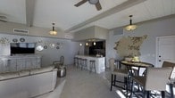 This single-floor space showcases an open-concept layout that seamlessly connects the living, dining, and kitchen areas into one cohesive and functional environment. The kitchen features a raised bar counter with stone-clad paneling, accommodating several bar stools, and is equipped with dark cabinetry and stainless steel appliances. Pendant lighting above the bar and a ceiling fan in the living area provide both practical illumination and stylistic detail. The living area is furnished with a plush neutral-toned sofa and an accent chair, complemented by a decorative metallic side table. An eclectic gallery of circular mirror and sunburst wall ornaments adds artistic character to the living space. The dining area is anchored by a dark bistro-style high table and upholstered high-back chairs, adjacent to a large textured wall accent that serves as a striking focal point. Light-toned wood-look flooring runs throughout, enhancing the sense of openness and continuity across the space. Neutral gray walls and recessed HVAC vents contribute to a clean, modern aesthetic. The overall ambiance is contemporary and well-curated, blending functional design with decorative flair.