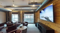 This single-floor lounge space blends industrial and rustic design elements to create a comfortable, visually engaging environment. Exposed silver ductwork runs prominently along the ceiling, lending an industrial character that contrasts beautifully with the warm, reclaimed wood-paneled accent wall anchoring one side of the room. Pendant lighting and recessed fixtures provide layered illumination throughout the space.

A large flat-screen television is mounted on the wood-paneled wall, making this area well-suited for entertainment and social gatherings. The seating arrangement features rich, dark brown leather club chairs grouped around a round wooden coffee table, offering an inviting and relaxed atmosphere. Patterned curtains frame the expansive windows that line the opposite wall, allowing generous natural light to fill the room while providing views of the surrounding urban streetscape.

The flooring consists of neutral carpeting that softens the overall aesthetic and adds comfort underfoot. A dark media console beneath the television provides practical storage. Together, the combination of industrial, rustic, and lounge-style elements gives this space a distinctive, well-curated ambiance ideal for relaxation and communal use.