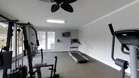 This single-floor fitness space offers a clean, functional environment designed to support a variety of workout routines. The room features neutral gray walls, white crown molding, and a smooth, light-colored ceiling that contribute to a bright and tidy atmosphere. A ceiling fan with dark blades is centrally mounted overhead, providing airflow and comfort during exercise sessions.

The space is equipped with a treadmill positioned centrally, a multi-functional weight and resistance training machine along one side, and a stationary bike, offering cardio and strength training options within a single room. A wall-mounted flat-screen television adds an entertainment element for users during workouts.

Natural light enters through a window along one wall, offering a view of the surrounding outdoor area and contributing to an open, airy feel. Interlocking foam floor mats are placed beneath the equipment to provide cushioning and protection. Overhead fluorescent lighting ensures the room remains well-lit at all times. The overall design is straightforward and practical, prioritizing functionality while maintaining a clean, uncluttered aesthetic suited for focused physical activity.