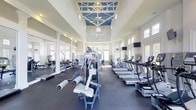 This fitness center offers a comprehensive workout environment designed to accommodate a wide range of exercise needs. The space is defined by its striking architectural ceiling, which features a decorative blue-and-white coffered design adorned with ceiling fans and a central ornamental medallion, lending the room an elevated, resort-style aesthetic. Soaring ceilings create an open, airy atmosphere throughout.

Natural light pours in through an abundance of large windows lining the perimeter walls, keeping the space bright and inviting. The flooring consists of durable dark rubber gym tiles, providing comfort and traction during workouts.

The cardio section is well-stocked with a row of treadmills and additional aerobic machines, while the strength training area features a selection of cable machines, weight benches, and free weights neatly organized along a mirrored wall. The mirrored wall further enhances the sense of spaciousness while allowing users to monitor their form. Mounted flat-screen televisions are positioned for easy viewing during cardio sessions. The overall layout is thoughtfully arranged to provide ample space between equipment, ensuring a comfortable and functional experience for all users.