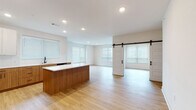 This contemporary single-floor residence offers a clean, modern aesthetic defined by a neutral palette, light hardwood flooring, and an abundance of natural light throughout. The open-concept living and kitchen area serves as the heart of the home, anchored by a substantial kitchen island with a crisp white quartz countertop and warm wood cabinetry. The kitchen is fully equipped with stainless steel appliances, including a French door refrigerator, electric range, and microwave, complemented by dual-tone cabinetry — white upper cabinets paired with natural wood lowers — and a matte black sink fixture. A striking black-hardware sliding barn door adds a stylish architectural accent to the living area, which opens to additional light-filled rooms with large windows dressed in horizontal blinds. The bedrooms feature plush carpet flooring and walk-in closets with built-in shelving. A secondary bedroom opens to an exterior door, providing convenient access. The primary bathroom offers a generous dual-sink vanity with wood cabinetry and a wide mirror, while a second full bathroom includes a tub-shower combo with built-in shelving. Recessed lighting is consistent throughout, contributing to the home's bright, polished atmosphere.