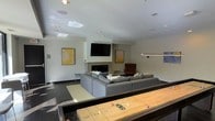 This contemporary single-floor space is thoughtfully designed for both relaxation and entertainment. The main lounge area is anchored by a sleek, modern fireplace set into a clean white wall, creating a warm and inviting focal point. A large wall-mounted flat-screen TV is positioned above the fireplace, making it ideal for gatherings and screenings. The seating arrangement features a generous sectional sofa in neutral gray, complemented by a plush area rug that defines the lounge zone. A classic mid-century accent chair in deep brown adds a stylish contrast to the otherwise cool-toned palette. The walls are adorned with abstract yellow artwork that introduces warmth and visual interest to the space. Recessed ceiling lighting provides even, ambient illumination throughout, while a sleek pendant light fixture above the game area adds a modern touch. A full-size shuffleboard table takes center stage in the foreground, offering a standout recreational feature that sets this space apart. The dark flooring grounds the room and creates a striking contrast with the bright white walls and ceiling. Overall, the space strikes a refined balance between comfort and play, making it well-suited for social events and casual everyday use.