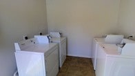 This single-floor laundry room is designed for practical, high-capacity use, featuring multiple sets of top-loading washers and dryers arranged along the walls. The room provides ample space to move between machines comfortably, making it well-suited for shared or high-demand laundry needs. The flooring consists of durable, earth-toned tile in a stone-like pattern, offering both resilience and ease of cleaning in a high-traffic environment. The walls are finished in a neutral, light gray tone that keeps the space feeling clean and functional. White baseboards provide a crisp contrast against the tiled floor. The machines are white, contributing to the room's clean, utilitarian aesthetic. The layout allows for efficient workflow, with machines positioned on opposing sides of the room, maximizing the use of available wall space. The overall design prioritizes functionality, providing a straightforward and organized environment for laundry tasks. The neutral color palette and simple finishes keep the focus on the room's practical purpose.