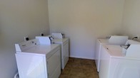 This single-floor laundry room is designed for practical, high-capacity use, featuring multiple sets of top-loading washers and dryers arranged along the walls. The room provides ample space to move between machines comfortably, making it well-suited for shared or high-demand laundry needs. The flooring consists of durable, earth-toned tile in a stone-like pattern, offering both resilience and ease of cleaning in a high-traffic environment. The walls are finished in a neutral, light gray tone that keeps the space feeling clean and functional. White baseboards provide a crisp contrast against the tiled floor. The machines are white, contributing to the room's clean, utilitarian aesthetic. The layout allows for efficient workflow, with machines positioned on opposing sides of the room, maximizing the use of available wall space. The overall design prioritizes functionality, providing a straightforward and organized environment for laundry tasks. The neutral color palette and simple finishes keep the focus on the room's practical purpose.