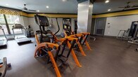Located at City Walk in Charlottesville, Virginia, this well-appointed fitness center offers a comprehensive workout environment spread across a single, thoughtfully organized floor. The space is defined by a bold and energetic design palette — gray walls accented by a distinctive yellow horizontal band near the ceiling — lending the facility a modern, motivating atmosphere throughout.

The cardio zone houses a row of premium Cybex elliptical trainers and additional aerobic machines, all finished in the brand's signature orange, set atop durable rubber flooring ideal for high-traffic exercise areas. A dedicated strength training section features cable machines, free weights, and weight benches arranged alongside floor-to-ceiling mirrors that enhance the sense of space and allow for proper form monitoring.

A separate group exercise studio provides a bright, inviting environment with hardwood-style flooring, large windows that invite natural light, and ceiling fans for added comfort. This area is equipped with spin bikes, TRX suspension trainers, stability balls, and a BOSU balance trainer, making it well-suited for diverse fitness classes. An adjoining functional training room offers additional specialty equipment, open floor space, and built-in storage shelving.

Recessed lighting throughout ensures consistent illumination, while a water fountain and accessible restroom facilities add convenience. Large glass entry doors and interior windows connect the various zones visually, creating a cohesive and welcoming fitness destination.