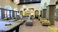 This single-floor space presents an expansive, elegantly appointed lounge area defined by its dramatic architectural character. The room features soaring ceilings accented by dark exposed wood beams, creating a striking contrast against the light-toned walls. Arched doorways and recessed niche details lend a Mediterranean-inspired aesthetic throughout the space.

At the heart of the room, a decorative fireplace serves as a focal point, framed by warm golden tile work and flanked by arched alcoves with subtle ambient lighting. The flooring consists of patterned Moroccan-style tiles in a neutral tone, adding texture and visual interest underfoot.

Seating is thoughtfully arranged to encourage conversation and comfort, featuring large sectional sofas and coordinating armchairs upholstered in shades of gray and olive green, accented with patterned throw pillows. Geometric pendant light fixtures hang from the ceiling, providing both functional and decorative illumination.

The overall ambiance is refined yet welcoming, blending classic architectural details with contemporary furnishings to create a sophisticated communal gathering space suited for both quiet relaxation and social interaction.