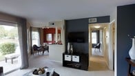 This single-floor residence offers a thoughtfully designed open-concept layout that connects the main living areas with ease and visual cohesion. The living room serves as the centerpiece of the home, anchored by a striking dark accent wall that provides a bold contrast against the light-toned carpet and neutral ceiling. A wall-mounted flat-screen television sits above a sleek black media console with built-in storage, adding both function and style to the space. The room opens naturally into a hallway leading to the private areas of the home, which are filled with natural light from well-placed windows. The dining area is positioned adjacent to the kitchen, defined by a warm red accent wall and illuminated by a modern track light fixture overhead. A breakfast bar with a white countertop separates the kitchen from the living and dining zones, providing a casual gathering space. The kitchen features dark countertops that complement the overall color palette. Throughout the residence, plush carpeting adds warmth and comfort underfoot, while multiple windows invite generous amounts of natural light, creating a bright and welcoming ambiance. The combination of bold color choices and contemporary furnishings gives this home a modern, energetic character.