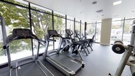 This fitness center is designed to provide a premium exercise experience within a well-appointed, single-level space. The room is defined by an expansive wall of floor-to-ceiling windows framed in sleek black mullions, flooding the interior with abundant natural light and offering open views of the surrounding urban streetscape, including mature trees and neighboring buildings. The generous glazing creates an airy, energizing atmosphere that enhances the workout environment.

The flooring consists of durable rubber gym matting in a dark tone, practical for both safety and equipment support. A row of commercial-grade Cybex treadmills is positioned along the window wall, allowing users to exercise while enjoying the outdoor views. Cybex upright bikes and additional cardio machines are arranged alongside the treadmills, creating an organized and efficient layout. The equipment is modern and well-maintained, reflecting a high standard of facility upkeep.

The ceiling is finished in white with integrated HVAC vents, ensuring comfortable climate control throughout the space. The overall design is clean, contemporary, and functional, with neutral tones and minimal ornamentation keeping the focus firmly on the fitness experience.