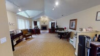 This single-floor facility is thoughtfully designed to serve both administrative and recreational needs. The leasing office area is a standout space, characterized by soaring vaulted ceilings that rise to a dramatic peak, drawing the eye upward and creating an open, airy atmosphere. A large angled window floods the room with natural light, complementing the track lighting overhead. Neutral carpet, warm-toned walls, and dark wood furnishings contribute to a professional yet welcoming environment. Multiple workstations are arranged throughout the room alongside seating areas, and a stone-surround fireplace serves as a central focal point, adding warmth and elegance. A round table with chairs provides an additional meeting space, while framed artwork and decorative accents complete the refined aesthetic.

The fitness center occupies a separate area of the building and is designed for comprehensive workouts. Floor-to-ceiling mirrors line the walls, visually expanding the space and allowing users to monitor their form. Large windows bring in natural light and outdoor views. The room is equipped with multiple treadmills, stair climbers, and a cable strength-training machine, all arranged on durable flooring. A wall-mounted television and ceiling fan add to the comfort and functionality of the space.