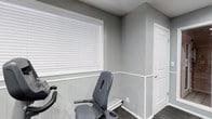 This single-floor space includes a fitness room finished with a refined two-tone wall treatment, featuring white shiplap-style wainscoting along the lower half and smooth gray-painted walls above, separated by a crisp white chair rail. The room is equipped with exercise equipment, including a stationary recumbent bike, and is finished with dark carpeted flooring that adds contrast to the lighter wall tones. A large window fitted with white horizontal blinds allows natural light to fill the space while offering privacy control. A white paneled door with a lever-style handle provides access to adjoining areas. The overall aesthetic is clean, neutral, and functional, with a cohesive gray and white color palette throughout. The space is well-suited for personal fitness use, combining practical design elements with polished architectural details that elevate the room beyond a standard utility space.