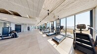 Located at 39 Bruton Street in San Francisco, CA, this expansive fitness facility delivers a well-appointed, single-floor layout designed for a comprehensive workout experience. The space is defined by dramatic floor-to-ceiling windows that line the exterior wall, flooding the interior with abundant natural light and framing sweeping views of the surrounding waterfront landscape and San Francisco Bay beyond.

The facility is organized across several distinct zones, accommodating a wide variety of training modalities. The cardio zone features a long row of treadmills, ellipticals, stationary bikes, and rowing machines, all positioned to take full advantage of the scenic outdoor views. Adjacent areas house an extensive selection of strength training equipment, including cable machines, multi-functional cable towers, weight benches, and selectorized machines suited for both upper and lower body training.

Architecturally, the space features a textured concrete ceiling with exposed structural elements and sleek pendant lighting throughout. Large-format tile flooring, concrete columns, and full-wall mirrors contribute to a modern, industrial aesthetic while enhancing the sense of openness. Tasteful potted plants are strategically placed throughout, adding a natural element to the contemporary design. The overall ambiance is bright, spacious, and energizing — a well-equipped facility suited for a broad range of fitness needs.