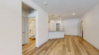 This 1-bedroom, 1-bathroom residence offers a clean, modern aesthetic defined by bright white walls, light-toned hardwood-style flooring, and recessed lighting throughout the main living areas. The open-concept kitchen and living room create a seamless, airy flow ideal for everyday living. The kitchen is outfitted with white shaker cabinetry, stainless steel appliances — including a refrigerator, range, and microwave — a subway tile backsplash, and a granite-topped peninsula with pendant lighting overhead, providing both prep space and a casual dining option. A built-in shelving nook adds a functional decorative touch nearby. The living area is spacious and bright, benefiting from natural light through a large front window with a transom above the entry door, complemented by a ceiling fan. The bedroom is carpeted for comfort, features its own ceiling fan, and includes a walk-in closet with built-in shelving. A second closet provides additional storage. The bathroom is well-finished with marble-style tile surrounding the tub/shower combo, a granite vanity, white cabinetry, and gold-toned fixtures. An in-unit laundry area is also present. This residence combines practical design with quality finishes throughout.