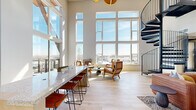 This sophisticated two-floor space is defined by its dramatic double-height ceilings and expansive floor-to-ceiling windows that flood the interior with natural light and frame sweeping outdoor views. The main level serves as the social hub, featuring an open-plan layout with light hardwood flooring throughout. A granite-topped bar counter with bar seating anchors the kitchen area, complemented by white cabinetry and a textured stone accent wall. The living area is generously proportioned, accommodating multiple seating arrangements beneath soaring ceilings and bathed in abundant natural light from the surrounding glass walls.

A striking black spiral staircase serves as both a functional element and a sculptural focal point, leading to the upper level. The second floor overlooks the main level through a sleek railing, offering an elevated perspective of the space. This level features a bold design statement wall adorned with geometric moss panels and wood accents, alongside a sculptural gold chandelier that adds warmth and visual drama.

The space extends outdoors to a rooftop deck finished with rich wood tile, offering panoramic views of the surrounding landscape. The deck is equipped with lounge seating and dining areas, making it an exceptional outdoor entertaining space under an open sky.