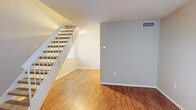 This two-story townhome is designed to maximize its compact footprint with a thoughtful, functional layout. The main level features an open living area finished with warm wood-look hardwood flooring that extends throughout. Neutral-toned walls in soft beige and greige create a clean, cohesive aesthetic, while a white-railed staircase with carpeted treads serves as a central architectural element. A window beneath the staircase allows natural light to filter into the lower level, brightening the space. Central air is provided via ceiling vents throughout.

Upstairs, the bedroom is generously proportioned for the overall scale of the home, featuring rich wood-look flooring, a ceiling fan with light fixture, and a standard reach-in closet with a hanging rod. Built-in shelving is also visible nearby, offering additional storage. A window overlooks the exterior, providing natural light and outdoor views. The upper level also includes a hallway landing area with its own window, adding to the home's brightness.

The bathroom includes tile flooring, a soaking bathtub with shower fixtures, a toilet, and a vanity topped with granite-style countertop material. Clean white trim and neutral walls complete the space, maintaining the home's consistent, understated design throughout.