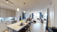 This single-floor residence offers a thoughtfully designed open-plan layout that seamlessly connects the kitchen, dining, and living areas. Light wood-style flooring flows throughout, complementing the clean white walls and contemporary finishes that define the space.

The kitchen is well-appointed with gray shaker-style cabinetry extending to the ceiling, stainless steel appliances including a French door refrigerator and range, white quartz countertops, a white subway tile backsplash, and a generous island with seating. Track and pendant lighting illuminate the workspace effectively.

The open living and dining area benefits from floor-to-ceiling windows and sliding glass doors that flood the space with natural light and offer expansive city views. A tan leather sofa, layered rugs, and bold large-format black-and-white photography create a curated, gallery-like atmosphere.

The bedroom continues the sophisticated aesthetic with a dark accent wall, oversized artwork, warm amber accent pillows, and direct access to a private balcony showcasing city skyline views.

The bathroom features a white vanity with an undermount sink, a glass-enclosed walk-in shower, and light tile flooring. A hallway bar cart and smart thermostat add practical, modern convenience throughout.