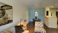 This single-story space showcases a cohesive interior design with rich, dark hardwood-style flooring that runs continuously throughout, creating a unified and grounded aesthetic. The living area is anchored by a stylishly patterned accent chair with a mid-century modern wooden frame, complemented by a coordinating ottoman. A warm terra cotta accent wall adds a bold pop of color to the entryway or transitional area, where a decorative side table displays curated accessories, including a statement ampersand sculpture.

The space transitions smoothly into a combined living and dining area. The living zone features comfortable seating, an adjustable arc task lamp, and soft, neutral wall tones that create a relaxed ambiance. Vaulted or sloped ceilings add a sense of openness to this area. The dining space is defined by a light blue accent wall, providing a calm contrast. A round dining table surrounded by bistro-style chairs sits beneath a simple pendant light, with a window nearby drawing in natural light. The overall design balances eclectic décor elements with a warm, inviting atmosphere throughout.