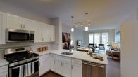 Located at 111 Morgan St in Stamford, Connecticut, this single-floor residence offers a thoughtfully designed open-concept layout that seamlessly integrates the kitchen, dining, and living areas into one cohesive living space. The kitchen is outfitted with crisp white shaker cabinetry, quartz countertops, and a full suite of stainless steel appliances, including a gas range, built-in microwave, and dishwasher. A peninsula with an undermount sink provides additional prep space and serves as a natural divider between the kitchen and the adjoining dining and living areas. Pendant lighting and a track light fixture illuminate the kitchen and transition zone, adding both function and a modern aesthetic touch. The living area extends toward large windows that invite generous amounts of natural light, creating a bright and welcoming ambiance throughout the open floor plan. The neutral color palette — soft grays, warm whites, and rich dark hardwood flooring — lends the space a clean, contemporary feel. The overall design reflects a refined urban sensibility, balancing practicality with modern style in a well-connected Stamford location.