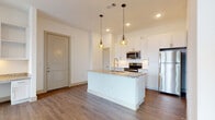 This single-floor residence offers a thoughtfully designed open-concept layout that seamlessly connects the kitchen and living areas. The kitchen is a standout feature, showcasing crisp white cabinetry extending to the ceiling, granite countertops, a central island with a built-in sink, and a full suite of stainless steel appliances including a range, over-the-range microwave, and refrigerator. A white subway tile backsplash and geometric pendant lights add a modern, polished touch. Warm wood-look flooring flows consistently throughout the space, complementing the clean white walls and bright recessed lighting. The living area is airy and open, benefiting from large windows that invite abundant natural light, with a ceiling fan adding both function and style. A hallway connects the living spaces to the bedroom and bathroom wing. The bathroom is elegantly finished with large-format marble-look tile in the walk-in shower, a frameless glass door, granite vanity countertop, and grey cabinetry offering ample storage. Adjoining the bedroom is a spacious walk-in closet equipped with double hanging rods and shelving, providing generous organizational capacity. The overall ambiance is modern, clean, and move-in ready.