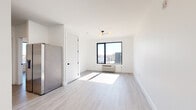 This thoughtfully designed single-floor unit showcases a bright, contemporary aesthetic with clean lines and a neutral palette carried consistently throughout. Light gray luxury vinyl plank flooring spans the main living and bedroom areas, complementing the crisp white walls and ceilings to create an airy, open feel.

The living area is bathed in natural light streaming through large black-framed windows that offer open views of the surrounding neighborhood. A modern multi-bulb ceiling fixture adds a stylish focal point to the room, while a built-in PTAC unit provides convenient climate control.

The kitchen is efficiently arranged along a single wall and features sage-gray shaker-style cabinetry extending to the upper walls for generous storage. Light quartz countertops provide ample workspace, and the suite of stainless steel appliances — including an electric range with oven, over-the-range microwave, and dishwasher — is complemented by a matte black single-handle faucet over a stainless steel sink.

The bathroom is finished with large-format gray tile flooring and features a wall-mounted white vanity with a matte black faucet, a framed mirror, and a clean, minimalist design. Black hardware accents on doors and fixtures tie the modern aesthetic together cohesively throughout the entire unit.