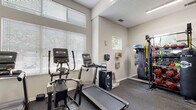 Located at 8531 Summersweet Ln in Raleigh, NC, this single-floor fitness center is thoughtfully designed to support a comprehensive workout experience. The main exercise area features durable speckled rubber flooring throughout, providing both safety and comfort during training sessions. Large windows fitted with horizontal blinds allow generous natural light to fill the space, creating a bright and energizing atmosphere. A ceiling fan overhead contributes to air circulation during intense workouts.

The cardio zone is equipped with multiple treadmills and an elliptical trainer, positioned to take advantage of the natural light from the windows. Opposite the cardio area, a full-length wall mirror expands the sense of space while allowing users to monitor their form. The strength training section is well-stocked with a tiered dumbbell rack, a power rack with barbell and weight plates, and additional resistance machines. A dedicated alcove houses a Matrix functional dual-cable trainer, ideal for versatile strength and rehabilitation exercises.

A separate room adjacent to the main floor serves as a stretch or activity area, featuring hardwood flooring and wall-mounted accessories. The common entry area includes a wall-mounted television, a mirror, and a waste receptacle. The facility's elevated ceilings enhance the open feel of the space, making it a practical and motivating environment for fitness enthusiasts of all levels.