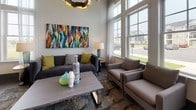 Located at 1600 Lake Seymour Dr in Middletown, Delaware, this single-story community clubhouse is designed with a sophisticated, contemporary aesthetic that balances style and functionality. The entrance foyer sets an immediate tone with dark wood-look tile flooring, an eye-catching geometric accent wall, and large frosted glass panels that invite natural light while maintaining privacy. A sleek barn door adds an industrial-modern touch to the transition between spaces.

The main lounge area is expansive and light-filled, boasting soaring double-height ceilings and floor-to-ceiling windows that frame views of the surrounding neighborhood and outdoor pool area. Multiple seating arrangements—featuring teal leather chairs, patterned sofas, and bold artwork—create distinct yet connected social zones. Wall-mounted flat-screen TVs serve as focal points in each lounge section.

A dedicated social kitchen and dining area features warm hardwood flooring, a long communal high-top table, modern pendant lighting, dark cabinetry, stainless steel appliances, and a full counter workspace with a sink—ideal for gatherings and events.

A quieter workspace nook offers a round table with seating, flanked by floor-to-ceiling green display shelving adorned with decorative vases, all bathed in natural light from oversized windows. Throughout, vibrant abstract artwork and curated décor elevate the overall ambiance of this well-appointed community space.