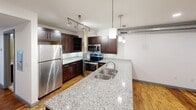 This two-floor loft in Charlotte's 28205 corridor showcases an industrial-modern aesthetic with exposed brick walls, wood-plank ceilings, and warm hardwood floors throughout. The main level offers generous open living space anchored by a gourmet kitchen featuring rich dark cabinetry, granite countertops, a large island with double sink, stainless steel appliances, and pendant lighting. Exposed ductwork and structural columns add authentic loft character. Rooms benefit from large windows—including a dramatic floor-to-ceiling brick-framed window—filling the space with natural light. Two full bathrooms are finished with granite vanities and tub-shower combos. The upper level provides a more intimate setting with clerestory windows. Throughout, grey trim and light hardwood floors tie the industrial and contemporary elements together seamlessly.