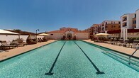 Located in Murrieta, California, this outdoor amenity area showcases a resort-caliber pool complex designed in a Spanish Colonial Revival style. The architecture features signature red clay tile roofs, white stucco facades, and arched detailing on the surrounding multi-story residential buildings, creating a cohesive and elegant aesthetic throughout the space.

At the heart of the area is a generously sized lap pool with clearly marked lanes, its crystal-clear turquoise water reflecting the abundant Southern California sunshine. Adjacent to the lap pool is a separate shallow wading or spa pool, providing additional relaxation options. The pool deck is finished with warm wood-toned porcelain tiles, offering a clean and modern contrast to the traditional architecture.

Lounge seating is thoughtfully arranged throughout the deck, including chaise lounges, stylish wicker egg swing chairs, market umbrellas for shade, and intimate seating groupings with accent chairs and tables. A dedicated clubhouse structure anchors one end of the pool area, featuring large glass panel openings that blur the boundary between indoor and outdoor spaces, with a mounted television visible inside.

The surrounding landscape opens to sweeping views of the hills and mountains characteristic of the Murrieta region, adding a scenic natural backdrop to the entire outdoor experience. The space is well-suited for both active recreation and leisurely relaxation.