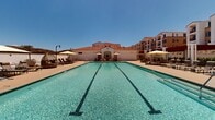 Located in Murrieta, California, this outdoor amenity area showcases a resort-caliber pool complex designed in a Spanish Colonial Revival style. The architecture features signature red clay tile roofs, white stucco facades, and arched detailing on the surrounding multi-story residential buildings, creating a cohesive and elegant aesthetic throughout the space.

At the heart of the area is a generously sized lap pool with clearly marked lanes, its crystal-clear turquoise water reflecting the abundant Southern California sunshine. Adjacent to the lap pool is a separate shallow wading or spa pool, providing additional relaxation options. The pool deck is finished with warm wood-toned porcelain tiles, offering a clean and modern contrast to the traditional architecture.

Lounge seating is thoughtfully arranged throughout the deck, including chaise lounges, stylish wicker egg swing chairs, market umbrellas for shade, and intimate seating groupings with accent chairs and tables. A dedicated clubhouse structure anchors one end of the pool area, featuring large glass panel openings that blur the boundary between indoor and outdoor spaces, with a mounted television visible inside.

The surrounding landscape opens to sweeping views of the hills and mountains characteristic of the Murrieta region, adding a scenic natural backdrop to the entire outdoor experience. The space is well-suited for both active recreation and leisurely relaxation.