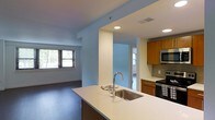 This single-floor residence showcases a thoughtfully designed open-concept layout that seamlessly connects the kitchen and living areas. The kitchen serves as a central hub, anchored by a large island with a sleek white countertop and an undermount stainless steel sink fitted with a contemporary arc faucet. Warm medium-toned wood cabinetry lines the walls, complemented by a neutral tile backsplash and stainless steel appliances, including a built-in microwave and electric range. Recessed lighting illuminates the space overhead, enhancing the clean, modern aesthetic. The kitchen flows naturally into the living area, which features rich dark hardwood flooring and large windows that invite generous natural light. Structural columns with crisp white finishes add architectural interest while subtly defining separate zones within the open floor plan. The overall ambiance is contemporary and functional, with a neutral color palette of whites and soft blues that creates a calm, welcoming atmosphere throughout the space.