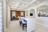 This single-floor space showcases a refined coastal aesthetic defined by consistent architectural detailing throughout. The main open area is anchored by striking coffered ceilings with recessed lighting, lending a sense of height and grandeur. A prominent marble-topped bar counter with blue chevron-upholstered barstools serves as a central gathering point, while a series of elegant arched openings connect the main area to adjacent spaces, including a glimpse into a well-appointed kitchen beyond.

The flooring throughout features a herringbone-patterned tile in a warm neutral tone, adding visual texture and cohesion across the space. A seating nook with large plantation-shuttered windows allows ample natural light and outdoor views, furnished with a blue plaid armchair for a welcoming touch.

A dedicated office area offers a rich wood built-in unit complete with cabinetry, open shelving, a built-in desk, and a murphy bed, maximizing both functionality and storage. Patterned drapery panels frame the windows consistently throughout, reinforcing the space's polished, coordinated design. The overall ambiance is bright, airy, and thoughtfully curated with a neutral and blue color palette.