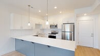 This two-story home offers a well-organized layout across two levels, with the main living areas on the lower floor and bedrooms occupying the more expansive upper floor. The entry level features an open-concept kitchen and living room finished with light hardwood-style flooring, recessed lighting, and a modern glass-panel interior door. The kitchen is a standout space, equipped with white shaker cabinetry, a white subway tile backsplash, quartz countertops, and stainless steel appliances including a refrigerator, range, and microwave. A two-tone island with a slate-blue base and white countertop provides additional workspace and serves as a natural divider between the kitchen and living area. The attached garage, accessible from the lower level, includes a concrete floor and an electrical panel. Upstairs, three bedrooms are finished with plush carpet flooring and neutral walls. The generously sized primary bedroom includes a dedicated walk-in closet with built-in shelving and hanging rods. Two full bathrooms serve the home, with one featuring light tile flooring, a white vanity, and clean, minimal finishes. A carpeted staircase connects both floors.