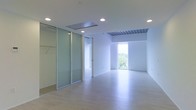 This single-floor space showcases a clean, minimalist aesthetic defined by an all-white interior palette and light-toned hardwood flooring that runs throughout. The open layout is enhanced by recessed ceiling lights that provide consistent, even illumination across the space. A notable architectural feature is the slatted ceiling detail visible in one section, adding subtle visual texture to an otherwise smooth, white ceiling plane.

Frosted glass sliding partitions divide portions of the space, offering flexible separation while maintaining a sense of openness and allowing light to pass through. These partitions include built-in closet rod infrastructure, suggesting dedicated storage functionality behind them. An adjacent doorway leads further into the space, hinting at additional rooms beyond the main area.

Natural light plays a significant role in the ambiance, with a large floor-to-ceiling window on the far wall framing views of surrounding greenery and the outside environment. This window floods the space with daylight, contributing to the bright, airy atmosphere. Overall, the space presents a highly functional, contemporary environment with an emphasis on light, openness, and adaptable design.