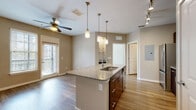 This well-appointed single-floor residence offers a thoughtfully designed open-concept layout that seamlessly connects the kitchen, dining, and living areas. The kitchen serves as the centerpiece of the space, anchored by a large central island topped with granite countertops and an undermount sink. Rich dark-espresso cabinetry — including glass-front upper cabinets — pairs elegantly with a mosaic tile backsplash and a full suite of stainless steel appliances, including a refrigerator, range, and over-the-range microwave. Pendant lighting above the island and a track lighting system overhead provide both task and ambient illumination throughout. The open living area features warm hardwood-style flooring, neutral taupe walls, and a ceiling fan that enhances comfort and airflow. A large window with blinds invites abundant natural light into the space. The bedroom is carpeted for comfort and features a pair of windows overlooking lush greenery, offering a tranquil outdoor view. The bathroom is finished with matching dark cabinetry, a granite-topped vanity, wood-look flooring, and a full shower-tub combination. An electrical panel is discretely accessible within the unit. Overall, the space presents a clean, contemporary aesthetic with cohesive finishes throughout.