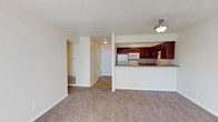 This single-floor residence offers a practical and welcoming layout designed for comfortable everyday living. The open-concept living area is finished with neutral-toned carpeting that adds warmth underfoot, while the light beige walls create a bright and airy atmosphere throughout the space. A half-wall partition with a raised breakfast bar separates the living area from the kitchen, maintaining an open feel while defining each zone. The kitchen is outfitted with rich, dark wood cabinetry that provides generous storage, complemented by stone-look countertops and a suite of white appliances including a refrigerator, microwave, and range. Overhead lighting in both the kitchen and the adjoining hallway ensures the space remains well-lit. A hallway extending from the main living area leads to additional rooms, with a door visible at its end suggesting private quarters or utility spaces beyond. A floor-level air vent is integrated into the baseboard near the living area, indicating a central HVAC system. The clean lines, neutral palette, and efficient floor plan make this a highly functional and move-in-ready space suited to a variety of lifestyles.