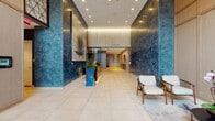 Located at 100 Sevilla Ave in the heart of Coral Gables, Florida, this ground-floor lobby presents a refined and architecturally distinctive environment designed to make a lasting first impression. The expansive entrance corridor is defined by dramatic deep-blue textured accent walls that contrast beautifully with warm wood paneling and large-format cream tile flooring, creating a sophisticated interplay of materials throughout the space.

At the center of the lobby, a sleek bronze reception desk is positioned beneath a striking large-scale botanical artwork, flanked by vibrant tropical plantings that reinforce the lush South Florida character of the space. Recessed and cove lighting throughout the lobby provides a warm, layered illumination that highlights the architectural details and finishes.

A comfortable seating area features contemporary wood-framed chairs and a round coffee table set atop an artistically designed area rug, offering a refined lounge setting for visitors. Glass-partitioned office space along one corridor provides a transparent and professional enclave with its own workstation and seating.

The elevator bank corridor, finished with textured neutral wall panels and elegant pendant lighting, provides seamless vertical access. Throughout, the design balances modern minimalism with warm, hospitality-influenced touches, resulting in a polished and welcoming atmosphere befitting its prestigious Coral Gables address.