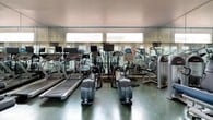 This fitness center is designed to support a full range of workout routines, combining thoughtful layout with a solid selection of equipment. The space is finished with polished green terrazzo-style flooring that adds a clean, professional aesthetic throughout.

The cardio zone is well-appointed with multiple treadmills and elliptical machines, each equipped with individual monitors to keep users engaged during their sessions. Strength training is equally well-served, with a variety of Star Trac selectorized weight machines targeting different muscle groups, along with free weights and a stability ball for more versatile training options.

A continuous wall of floor-to-ceiling mirrors runs along one side of the room, visually expanding the space and allowing users to monitor their form. Track lighting is mounted along the ceiling, providing bright, even illumination throughout. A wall-mounted flat-screen television adds an entertainment element to workouts.

Large windows along the perimeter flood the space with natural light, while green-framed French doors open directly to an outdoor patio area, offering a seamless connection to the exterior. The overall ambiance is bright, functional, and welcoming, making it a practical and motivating environment for fitness.
