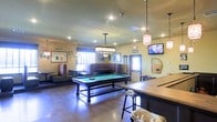 This entertaining pub-style space is designed for social gatherings and recreation, offering a well-appointed single-floor layout filled with engaging amenities. At the heart of the room sits a full-size pool table with a vibrant green felt surface, illuminated by a rectangular pendant light fixture that draws the eye and anchors the space. To one side, a long shuffleboard table provides additional game-play options, accompanied by upholstered bar stools with a classic turned-leg design. Comfortable dark leather booth seating lines one wall, offering a relaxed lounging area ideal for conversation or watching the wall-mounted flat-screen TVs positioned throughout the room. The warm yellow walls contribute to an inviting, pub-like ambiance, while decorative touches — including an Irish-themed sign and framed artwork — add character and personality. Pendant lantern-style light fixtures hang from the ceiling alongside recessed lighting, ensuring the space is well-lit throughout. Polished concrete floors provide a durable, low-maintenance surface that complements the casual yet spirited aesthetic. Natural light filters in through windows with blinds, adding brightness to the overall atmosphere. This space is thoughtfully designed to serve as a versatile entertainment hub.